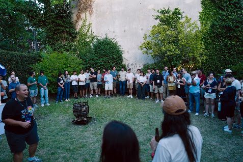 The whole Summer Camp 2026 group gathered in a garden around a fire pit at dusk