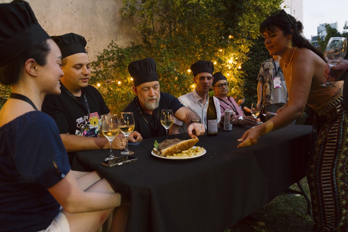 Campers in chef hats sharing an asado around a long table, lit by string lights