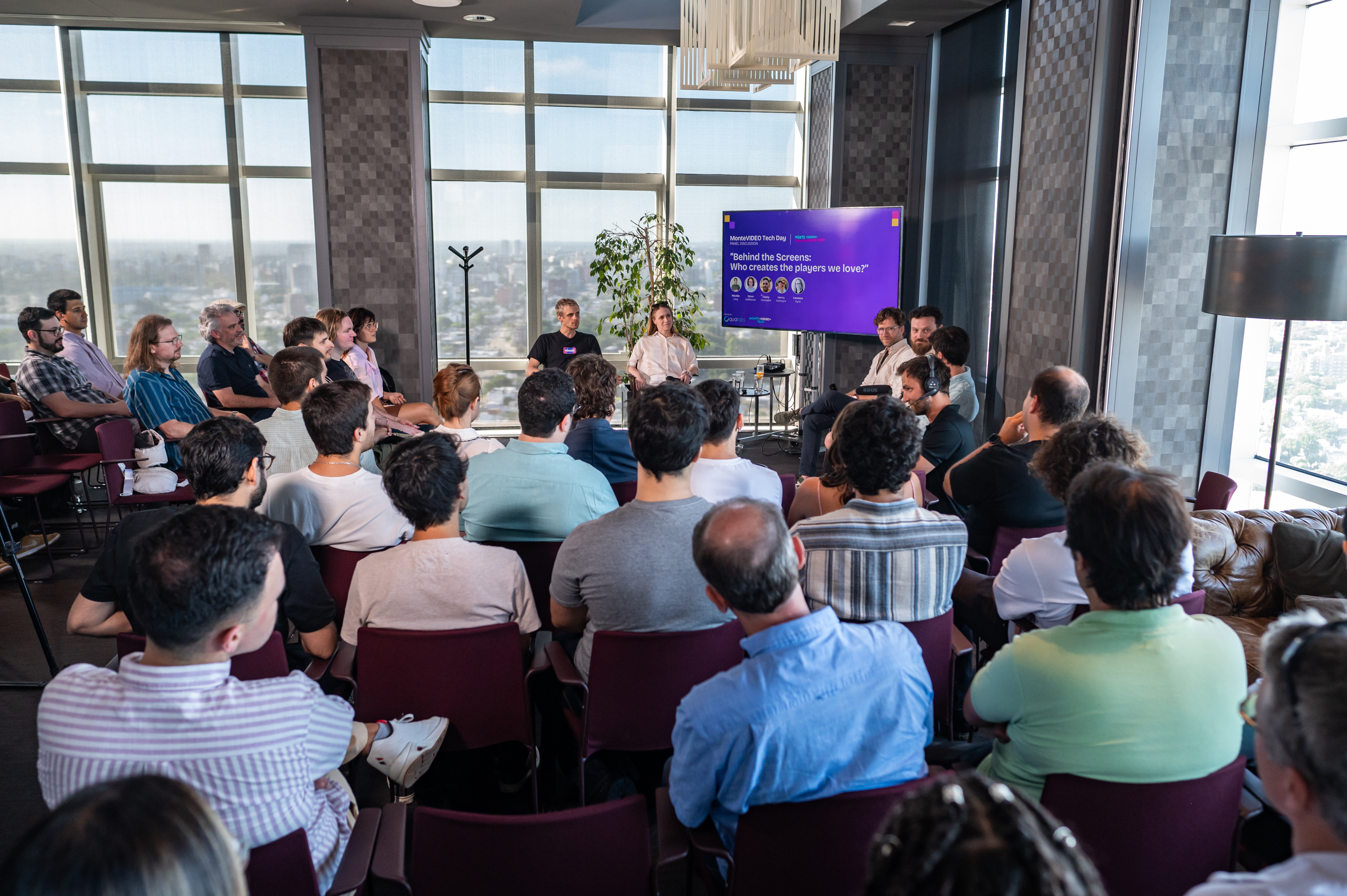 Full room of attendees watching the MonteVIDEO Tech Day panel, with the Montevideo skyline through the windows