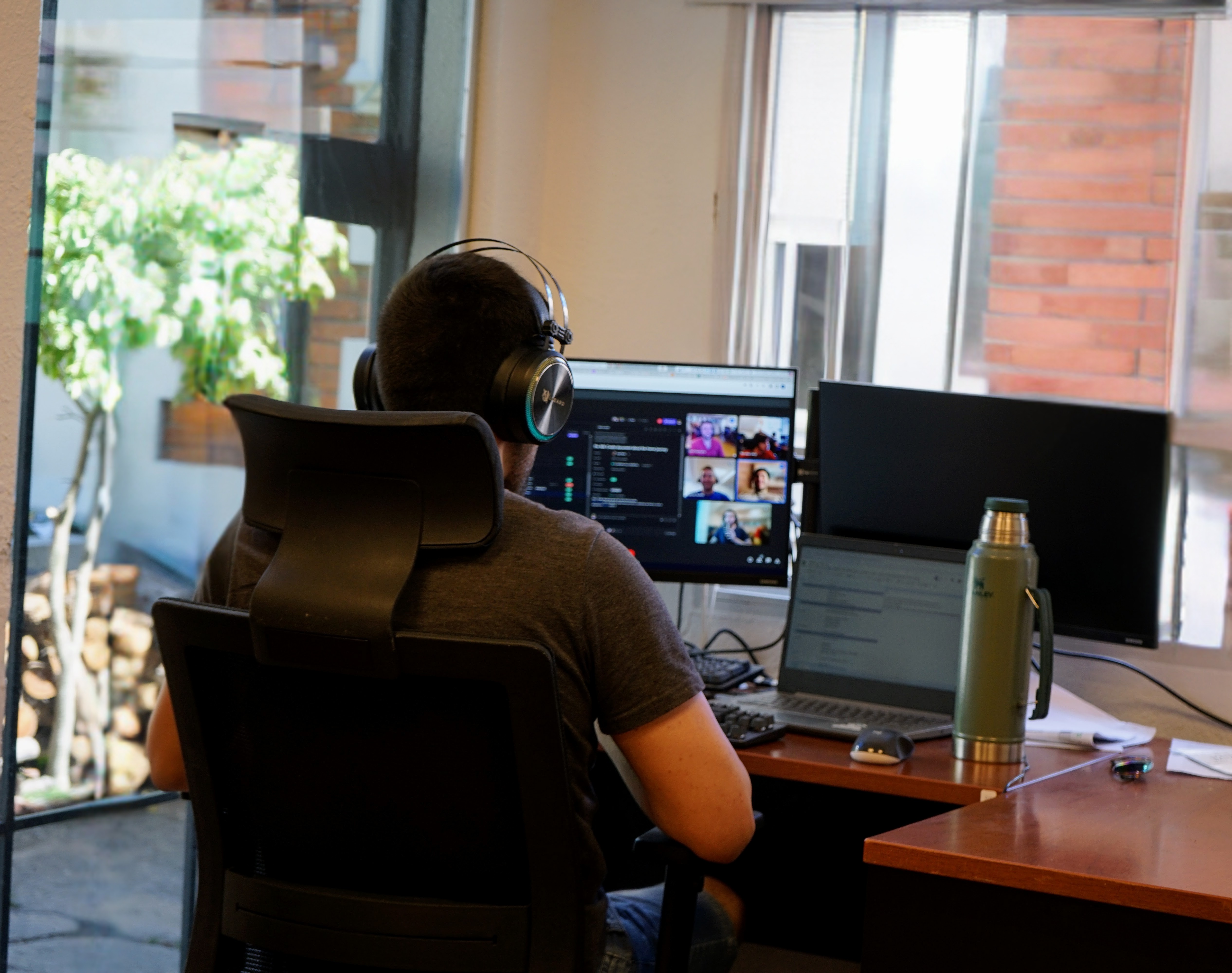 A participant working from a quiet Qualabs office desk, on a video call with two screens and a thermos of mate