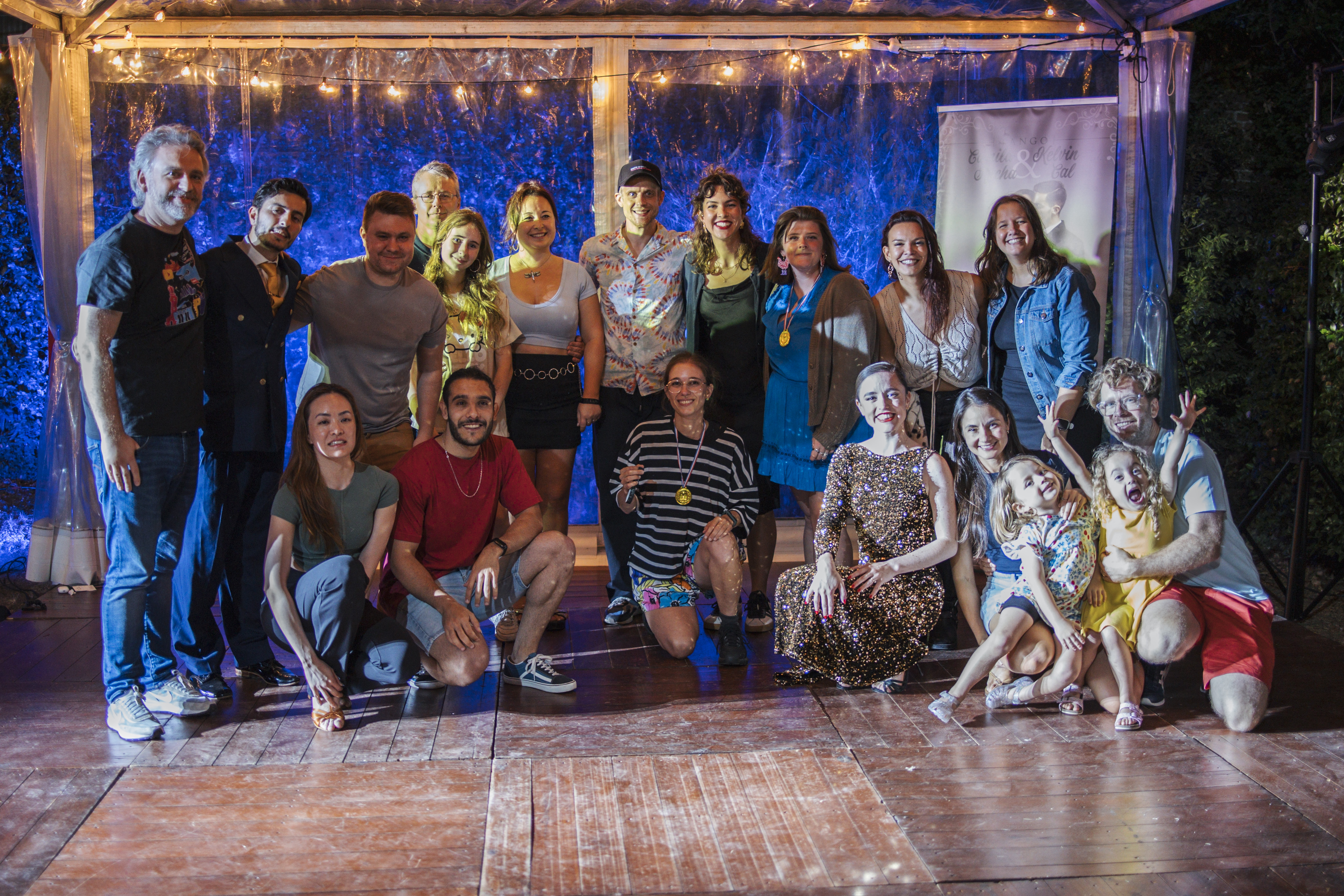 Group photo of Summer Camp participants and their families under a pergola strung with lights at the closing party