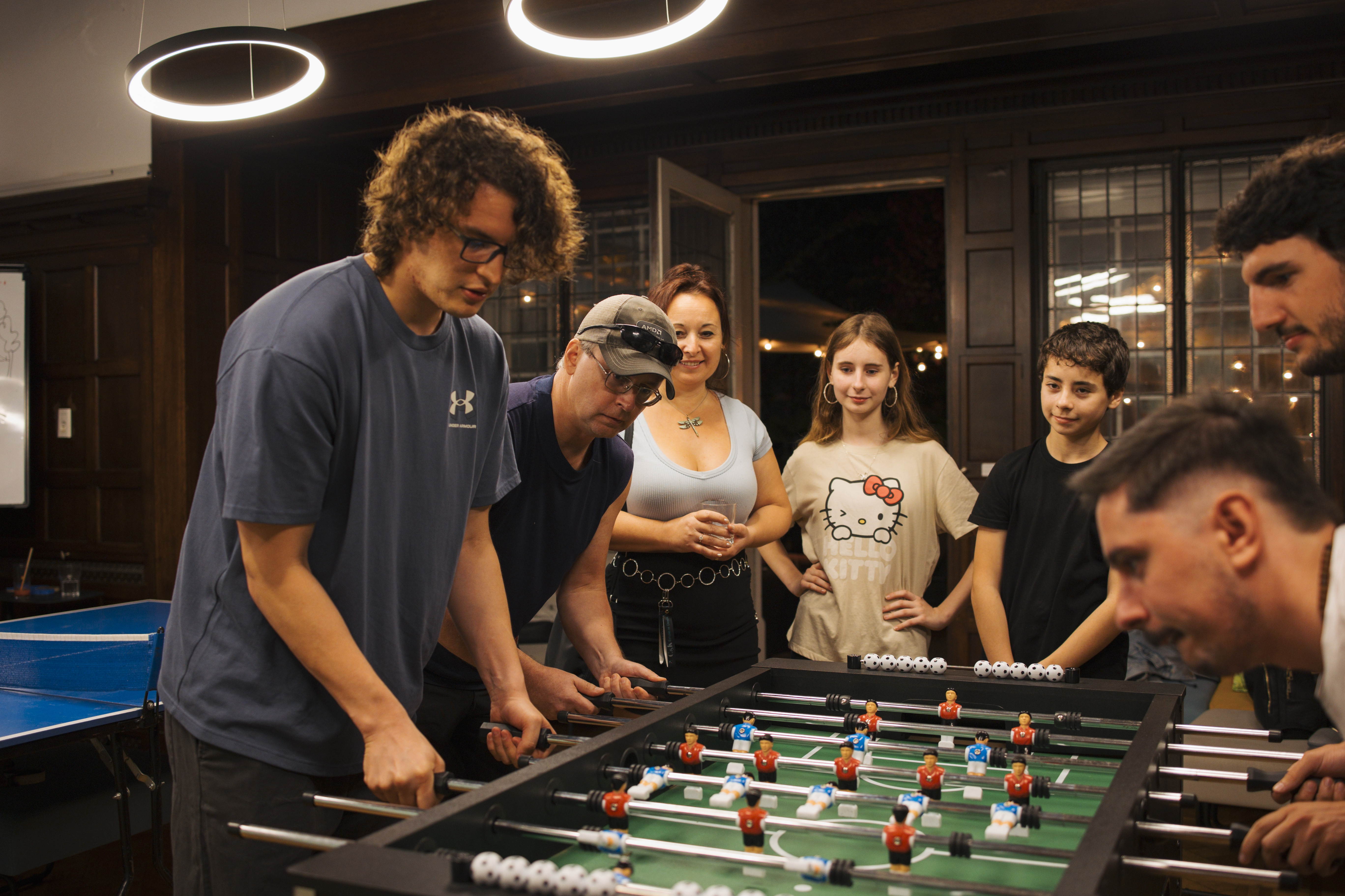 Participants and kids gathered around a foosball table during a Summer Week social night