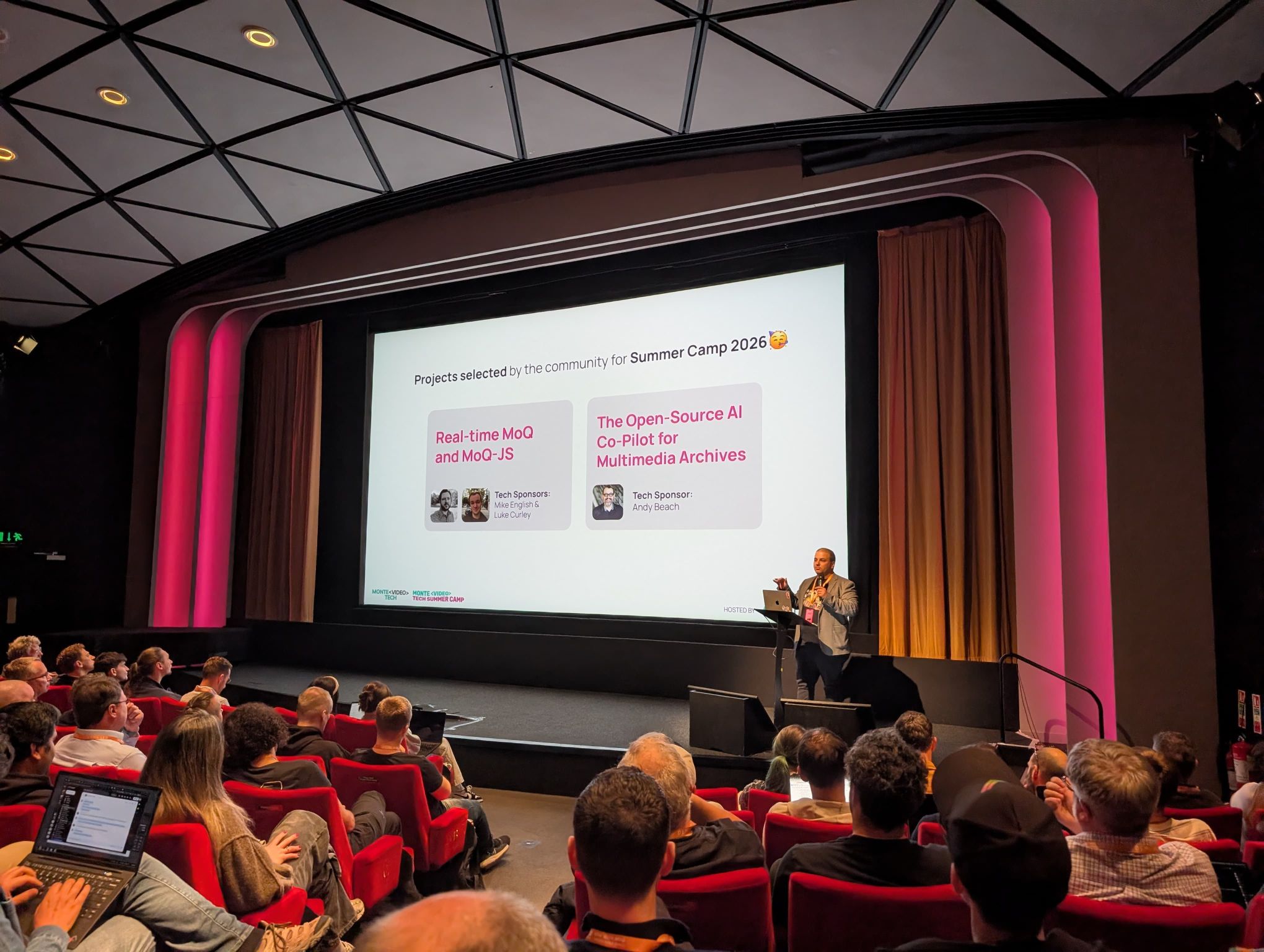 A speaker on stage at Demuxed presenting the slide “Projects selected by the community for Summer Camp 2026”, with two projects: “Real-time MoQ and MoQ-JS” (Tech Sponsors: Mike English & Luke Curley) and “The Open-Source AI Co-Pilot for Multimedia Archives” (Tech Sponsor: Andy Beach)